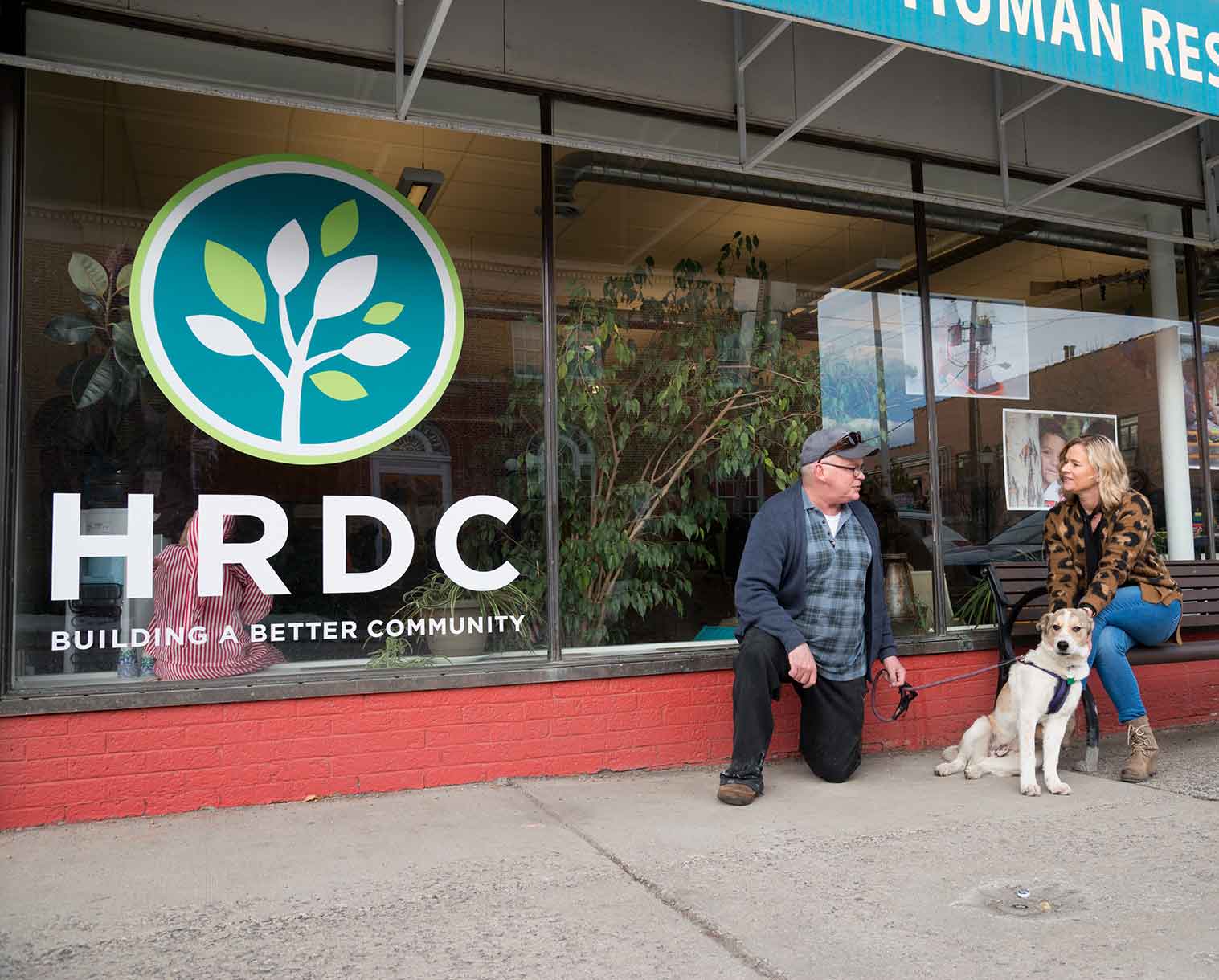 Photo of one person kneeling on the ground holding the leash to a dog that is sitting nearby. Another person is sitting on a bench. The two are talking to one another in front of the HRDC office. The logo on the windows reads, HRDC Building a Better Community.