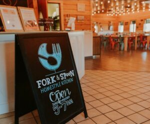 Photo of a sandwich board just inside of Fork and Spoon. Text on the board reads, Fork & Spoon, Homestyle Kitchen. Open Sunday - Friday, 5-7 pm. 